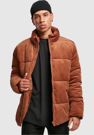Brun kordfløyels pufferjakke med quiltet design, høy krage og glidelås. Båret over en svart skjorte, som viser ribbet tekstur og polstring.