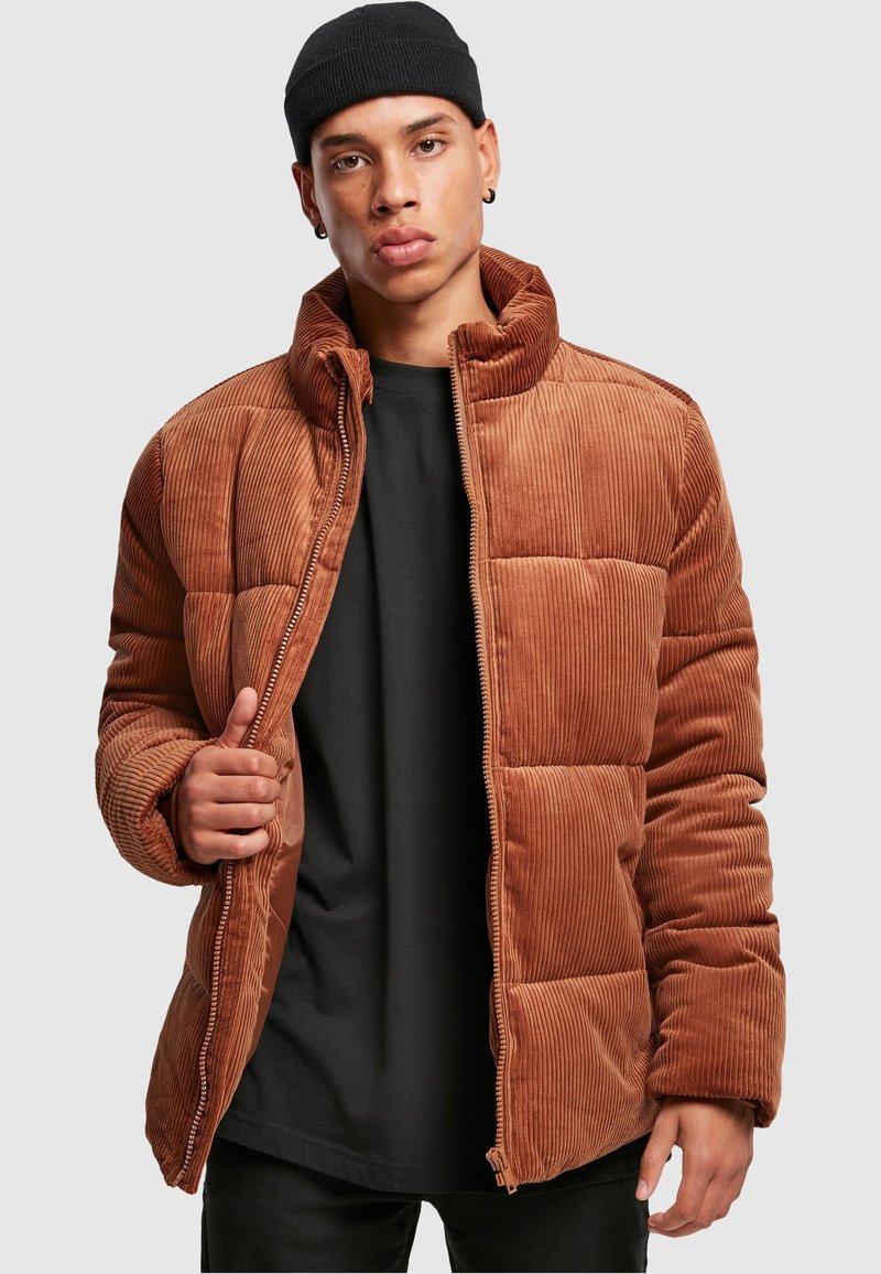 Brun kordfløyels pufferjakke med quiltet design, høy krage og glidelås. Båret over en svart skjorte, som viser ribbet tekstur og polstring.
