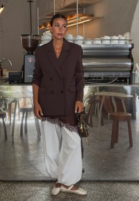 Brązowa marynarka dwurzędowa na białych luźnych spodniach z koronkowym wykończeniem. Srebrne baleriny i torebka w wzory. Minimalistyczne tło kawiarni.