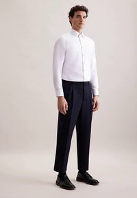 Biała koszula z długim rękawem i kołnierzykiem na guziki w połączeniu z ciemnoniebieskimi spodniami i czarnymi butami, prezentująca dopasowany krój i gładką tkaninę.
