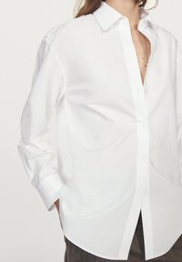 Camicia bianca in cotone con colletto, maniche lunghe e chiusura frontale con bottoni. Presenta una vestibilità rilassata e un orlo curvo. Texture liscia.