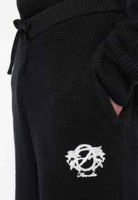 Czarne dzianinowe spodnie o fakturowanej powierzchni, z regulowanym pasem na sznurek i bocznymi kieszeniami. Charakterystyczne białe wyszywane logo po boku.