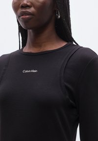 Juoda ilgomis rankovėmis suknelė su apvalia iškirpte ir balta Calvin Klein logotipu. Lygi, tampri medžiaga, prigludęs dizainas ir akcentinės siūlės.