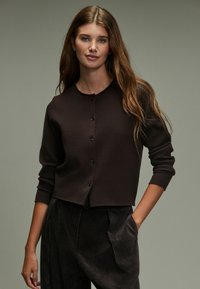Donkerbruine gebreide cardigan met een ronde halslijn, knopen aan de voorkant en lange mouwen. Draagt over een gestructureerde, donkerbruine corduroy broek.