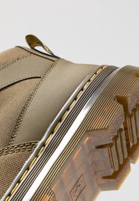 Acercamiento a la suela de una bota rústica de color bronceado con una plantilla translucida y texturizada, y costura amarilla a lo largo del borde de la parte superior de tela.