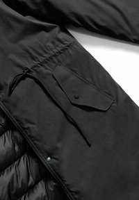 Giacca nera con un materiale esterno liscio, una texture trapuntata nella parte inferiore e una tasca con bottoni a pressione e cordoncino in vita.