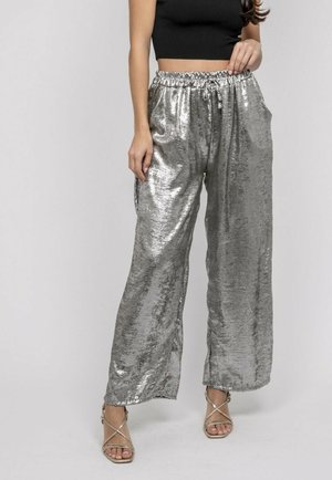 Femme portant un pantalon large en métal argenté avec une taille à cordon, un haut court noir, et des sandales dorées à lanières, sur fond gris.