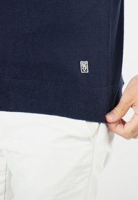 Pull en maille bleu marine avec un ourlet et des poignets côtelés ; présente une petite étiquette de logo blanche sur le bas à gauche. Porté sur un pantalon blanc.