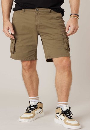 Homme portant un short cargo kaki, un t-shirt noir, des baskets haute en beige et blanc, et des bracelets de perles, debout devant un fond uni.