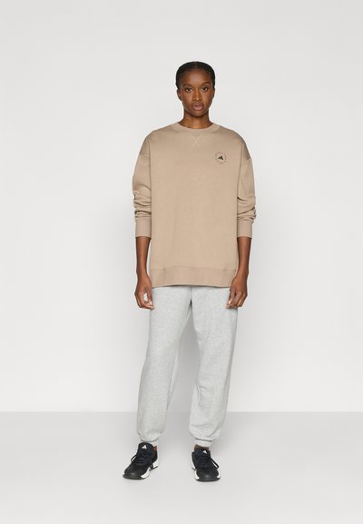 Ανοιχτό μπεζ φούτερ με στρογγυλή λαιμόκοψη και λεπτομέρεια με λογότυπο, συνδυασμένο με γκρι φόρμα και μαύρα αθλητικά παπούτσια με κορδόνια.