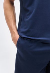 Αρσενικό χέρι που ακουμπά στην τσέπη σκούρων μπλε παντελονιών, φορώντας αντίστοιχο σκούρο μπλε πάνω μέρος, μπροστά σε απλό φωτεινό φόντο.