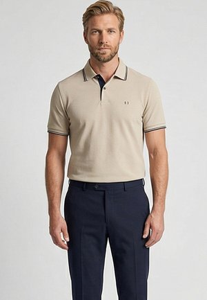 Man draagt een beige poloshirt met zwarte rand en marineblauwe broek, staat voor een effen lichte achtergrond en kijkt recht vooruit.