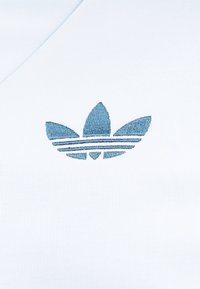 Modré vyšívané logo Adidas trefoil prišité na bielej látke, detailný pohľad na odevný detail.