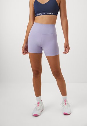 Femme portant un soutien-gorge de sport bleu marine, un short cycliste taille haute violet clair, des chaussettes blanches et des baskets blanches avec des touches roses.