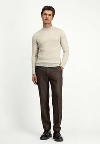 Beżowy sweter z dzianiny o wysokim kołnierzu i prążkowanym wykończeniu, zestawiony z brązowymi spodniami w kant. Modelka stoi z rękami w kieszeniach, nosząc czarne buty.
