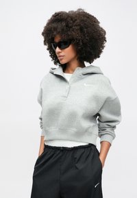 Γκρι κοντή φόρμα με μπροστινό κούμπωμα με κουμπιά και λογότυπο, συνδυασμένη με μαύρο φαρδύ παντελόνι. Το μοντέλο φοράει σκουρόχρωμα γυαλιά ηλίου, με εμφανή σγουρά μαλλιά.