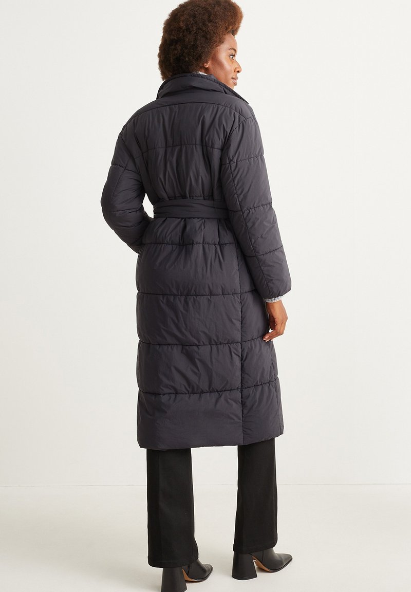 Langer, dunkler Daunenmantel mit tailliertem Gürtel. Verfügt über horizontale Nähte, einen hohen Kragen und seitliche Taschen. Trägt ihn über schwarzen Hosen und Stiefeln.