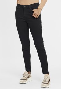 Fekete, magas derekú, szakadt szegélyű skinny farmernadrág elülső zsebekkel és gombos záródással. Vastag, bézs színű sportcipőkkel párosítva.