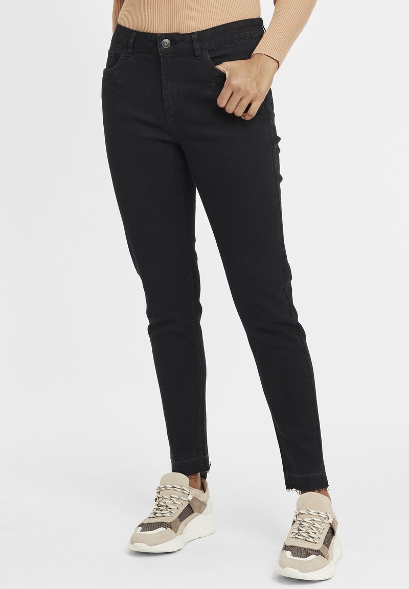 Fekete, magas derekú, szakadt szegélyű skinny farmernadrág elülső zsebekkel és gombos záródással. Vastag, bézs színű sportcipőkkel párosítva.