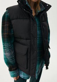 Czarna kamizelka puchowa z wysokim kołnierzem, zatrzaskami i boczną kieszenią, noszona na zielonej flanelowej koszuli w kratę o różnej fakturze.