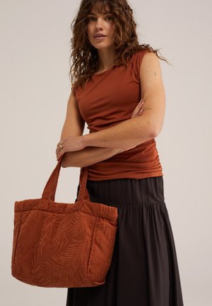 Frau mit lockigem braunem Haar trägt ein rostfarbenes ärmelloses Top und einen schwarzen Rock, hält eine große rostfarbene gemusterte Tragetasche.
