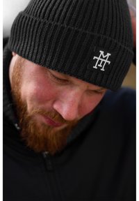 Black ribbed knit beanie with a sewn white logo patch. Worn slightly folded, showcasing texture and warmth. Person's face partially visible.