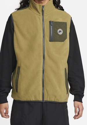 Personne portant un gilet polaire zippé vert olive avec des accents de poche vert foncé, sur un tee-shirt noir à manches longues et un pantalon noir.