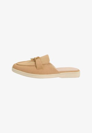 Mocassin beige en daim sans talon avec une petite boucle en métal doré et une semelle épaisse en caoutchouc blanc.