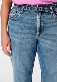 Femme portant un jean en denim bleu taille haute avec une ceinture en denim assortie et un haut rose rentré, main gauche reposant sur la hanche.