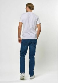 Bela bombažna majica, kratki rokavi, zadnji pogled. V kombinaciji z modrimi kavbojkami, ravnega kroja, s subtilnim šivanjem in standardnimi žepi.