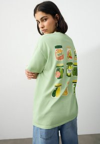 T-shirt en coton vert clair avec des graphismes colorés de bocaux au dos, représentant divers aliments en conserve. Coupe ample et décontractée avec des manches courtes.