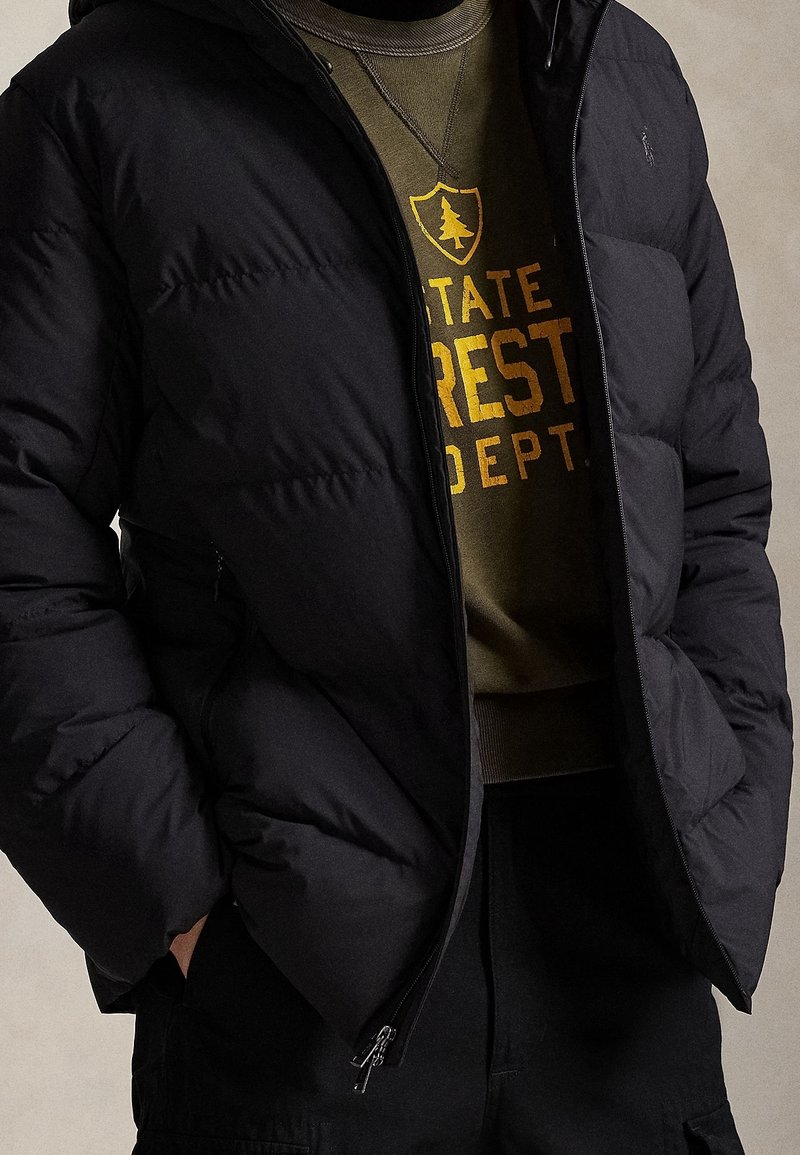Personne portant une doudoune noire sur un sweat vert olive avec le texte jaune « STATE FOREST DEPT » et un emblème d’arbre.