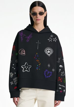 Personne portant un sweat à capuche noir avec des griffonnages colorés comprenant des étoiles, des cœurs, un crâne, des oiseaux et des symboles, associé à un pantalon beige et des lunettes de soleil ovales.