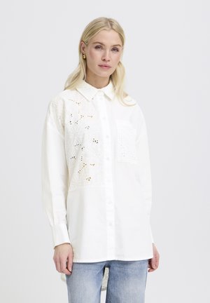 Woman with blonde hair wearing oversized white button-up shirt with floral cutout patterns and light blue jeans, standing against plain background.