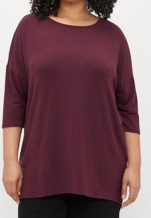 Frau trägt ein locker sitzendes, dunkelviolettes Dreiviertelarmshirt und schwarze Hose vor hellem, einfarbigem Hintergrund.