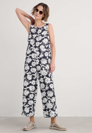 Schwarz-weißes Blumen-Overall mit weitem Bein, ärmellosem Design und rundem Ausschnitt, kombiniert mit beigefarbenen, strukturierten Schuhen.
