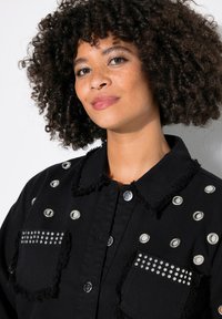 Zwarte spijkerjas met zilveren studded en oogjes, franjes op de kraag en zakken, met knoopsluiting en gestructureerde stof.