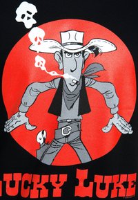 Mustat t-paidat, jossa on sarjakuvamuotoinen cowboy leveälierinen hattu, punainen huivi ja piippu, punaisella pyöreällä taustalla. Teksti kuuluu "LUCKY LUKE."