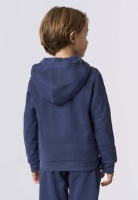 Bambino con capelli corti castani che indossa un felpa blu navy e pantaloni abbinati, girato di spalle contro uno sfondo chiaro e neutro.