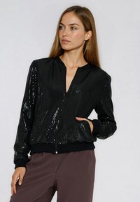 Czarna kurtka bomberka wykonana z cekinowej tkaniny z zamkiem błyskawicznym z przodu, ściągaczami na mankietach i u dołu oraz bocznymi kieszeniami. Gładka faktura z błyszczącymi akcentami.