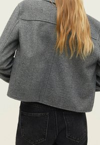 Szary żakiet w wzór w jodełkę o kroju cropped, charakteryzujący się gładką fakturą i obniżonymi ramionami. Widok z tyłu ukazuje dwie boczne kieszenie.