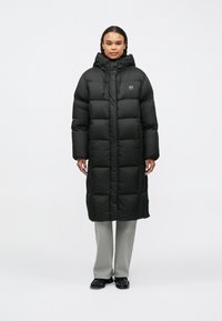 Mulher a usar um longo casaco acolchoado preto com capuz, calças cinzas e sapatos pretos, de pé contra um fundo claro e liso.