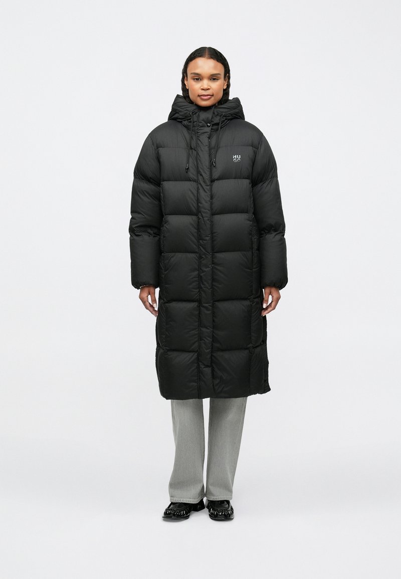 Mulher a usar um longo casaco acolchoado preto com capuz, calças cinzas e sapatos pretos, de pé contra um fundo claro e liso.