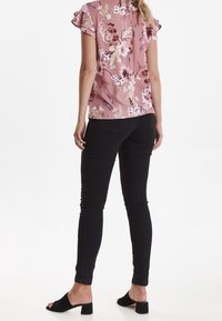 Blusa floreale rosa con maniche corte a volant, abbinata a jeans neri attillati. La modella indossa mule nere con punta scoperta. Lo sfondo è bianco uniforme.