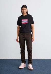 Black cotton t-shirt with colorful "PUMA" graphic, paired with brown pants and light pink sneakers featuring white laces and gum sole.
