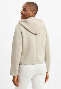 Beige Cropped-Hoodie mit Kapuze, strukturiertem Stoff und sichtbaren Nähten, kombiniert mit hellen Hosen.