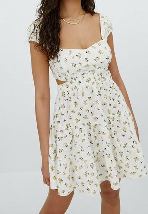 Vrouw draagt een witte jurk met korte mouwen met geel bloemenprint, geschulpte zoom en uitgesneden zijkanten, gecombineerd met gelaagde gouden kettingen.