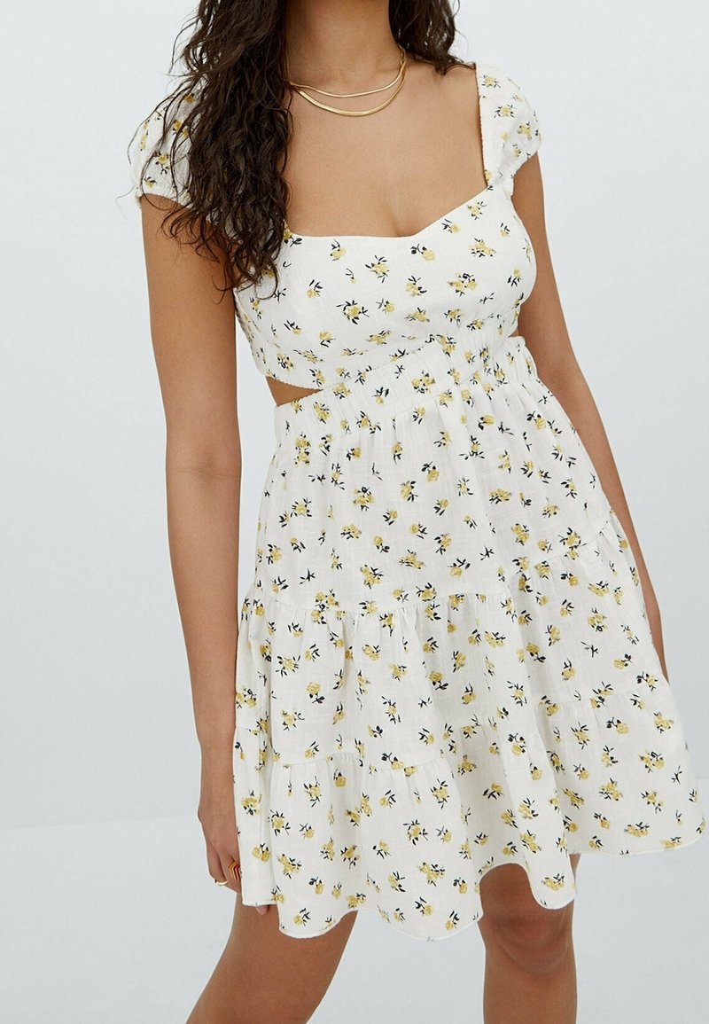 Femme portant une robe blanche à manches courtes avec un imprimé floral jaune, un ourlet festonné et des côtés découpés, accessoirisée de colliers dorés superposés.