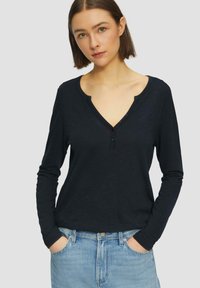 Dunkelblauses Langarmshirt mit V-Ausschnitt und einer Drei-Knopf-Leiste, kombiniert mit hellblauen Jeans. Weiche Textur und entspannte Passform.