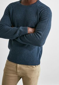 Marinblå stickad tröja med ribbad fåll och manschetter, med texturerade axelaccenter. Matchad med ljusbeige byxor.